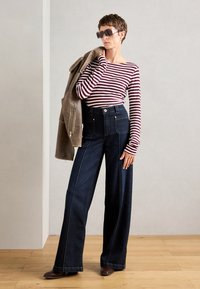 Stripet langermet topp, mørke vide jeans med frontlommer, og brune ankelstøvler; modellen holder en beige jakke og har solbriller på.