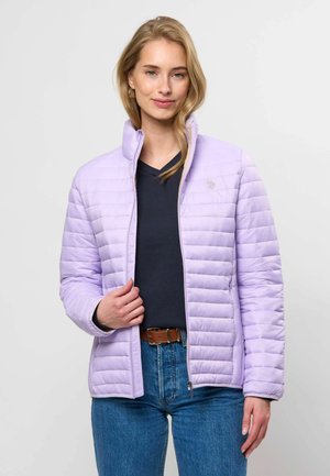 Kvinde iført lys lilla quiltet jakke over sort top og blå jeans, stående mod en ensfarvet hvid baggrund.