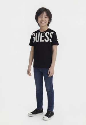 Bambino sorridente con capelli neri che indossa una maglietta nera "GUESS", jeans blu e scarpe da ginnastica nere, in piedi davanti a uno sfondo bianco uniforme.