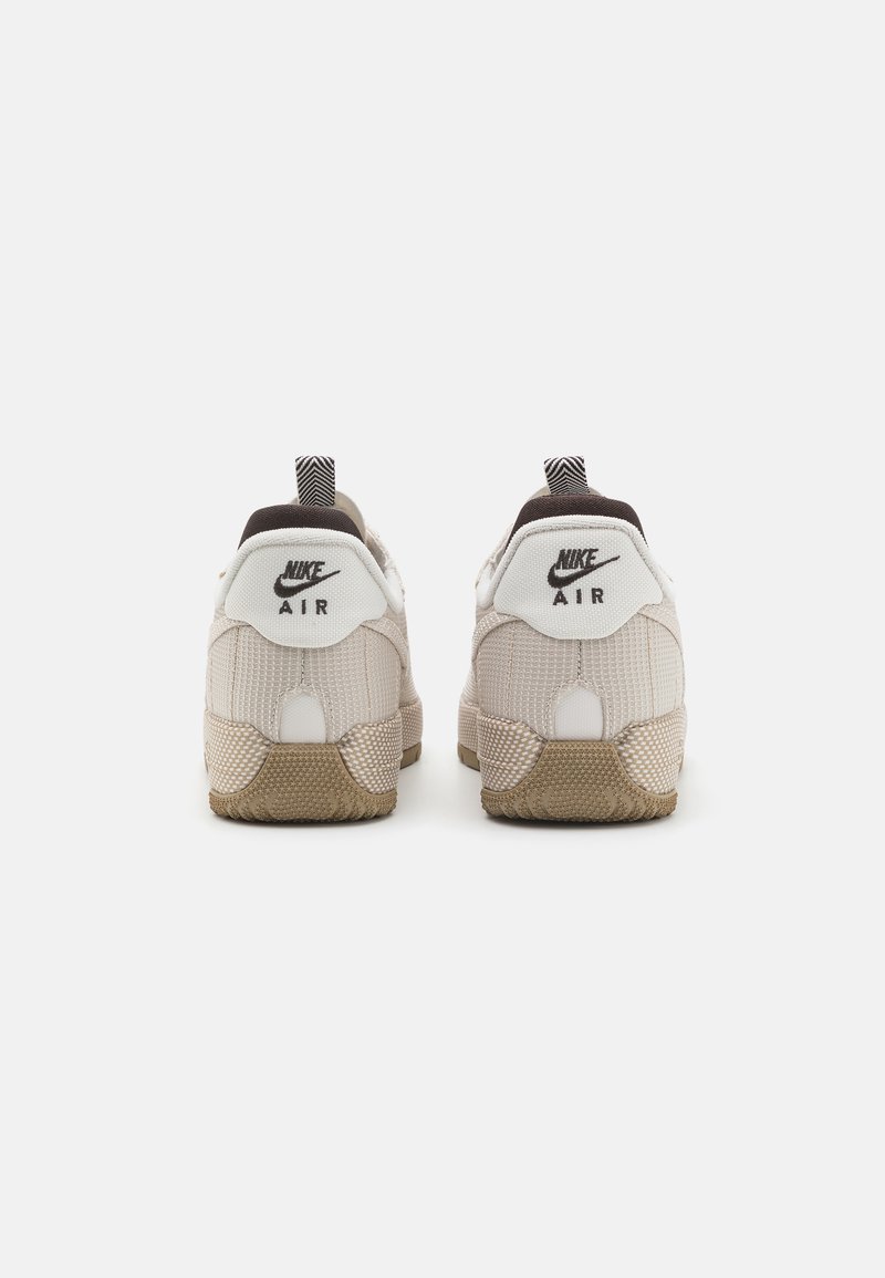 Une paire de baskets Nike Air beiges vue de l'arrière, avec des semelles texturées et des logos Nike noirs sur des panneaux blancs au niveau du talon.