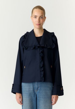 Femme portant une veste bleu marine avec un col à volants et des bordures, des poignets boutonnés, et un jean bleu clair, debout devant un fond blanc uni.
