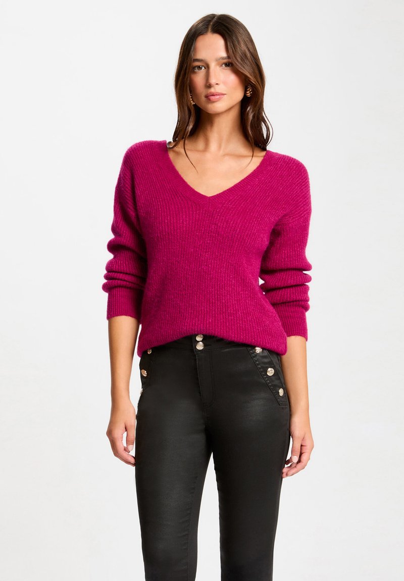Un pull en maille rose à col en V avec des manches longues, en tissu texturé, associé à un pantalon noir taille haute avec des boutons métalliques sur le côté.