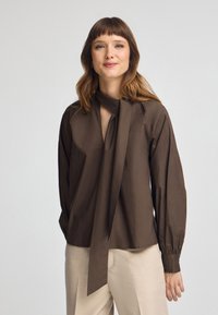 Blusa marrone a maniche lunghe con scollatura a V profonda e dettaglio a nodo al colletto. Caratterizzata da maniche arricciate e texture liscia, abbinata a pantaloni chiari.