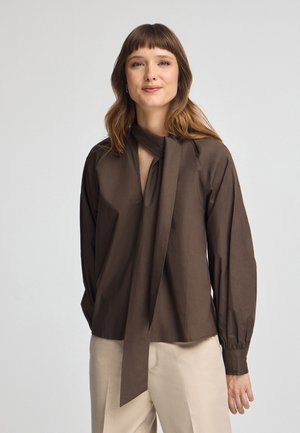 Blusa marrón de manga larga con escote en V profundo y detalle de lazo en el cuello. Presenta mangas recogidas y textura suave, combinada con pantalones claros.