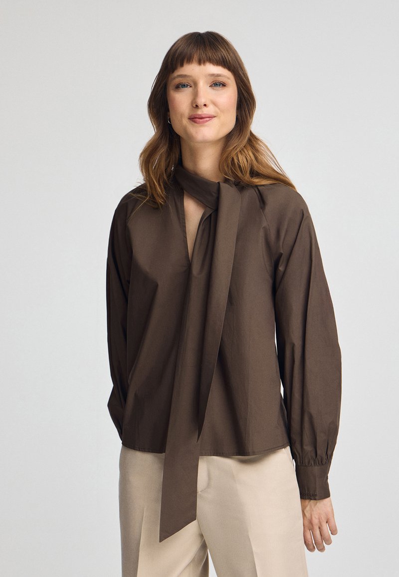 Blusa marrone a maniche lunghe con scollatura a V profonda e dettaglio a nodo al colletto. Caratterizzata da maniche arricciate e texture liscia, abbinata a pantaloni chiari.
