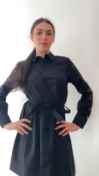 Kvinde med langt mørkt hår iført en sort skjortekjole med bælte og blondeærmer, stående med hænderne på hoften mod en hvid baggrund.