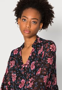 Blusa preta com padrão floral, apresentando um decote em V profundo, adornada com flores rosa e azuis em tecido transparente, e mangas largas e fluidas.
