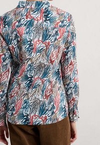 Blusa blanca con un patrón colorido de corales y algas marinas en azul, rojo y marrón. Mangas largas con puños abotonados; tejido suave y ligero.
