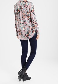 Camicia a maniche lunghe con motivo floreale, caratterizzata da una combinazione di rosa, verde e nero su uno sfondo chiaro, abbinata a jeans skinny blu scuro e stivali neri con tacco.
