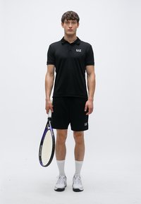 Svart poloskjorte med EAZ-logo, svarte shorts, holder en tennisracket med lilla detaljer, hvite ankelsokker og treningssko.