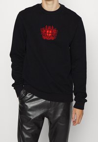 Man som bär en svart sweatshirt med röd broderad sköld och svarta läderbyxor, stående med ena handen i fickan.