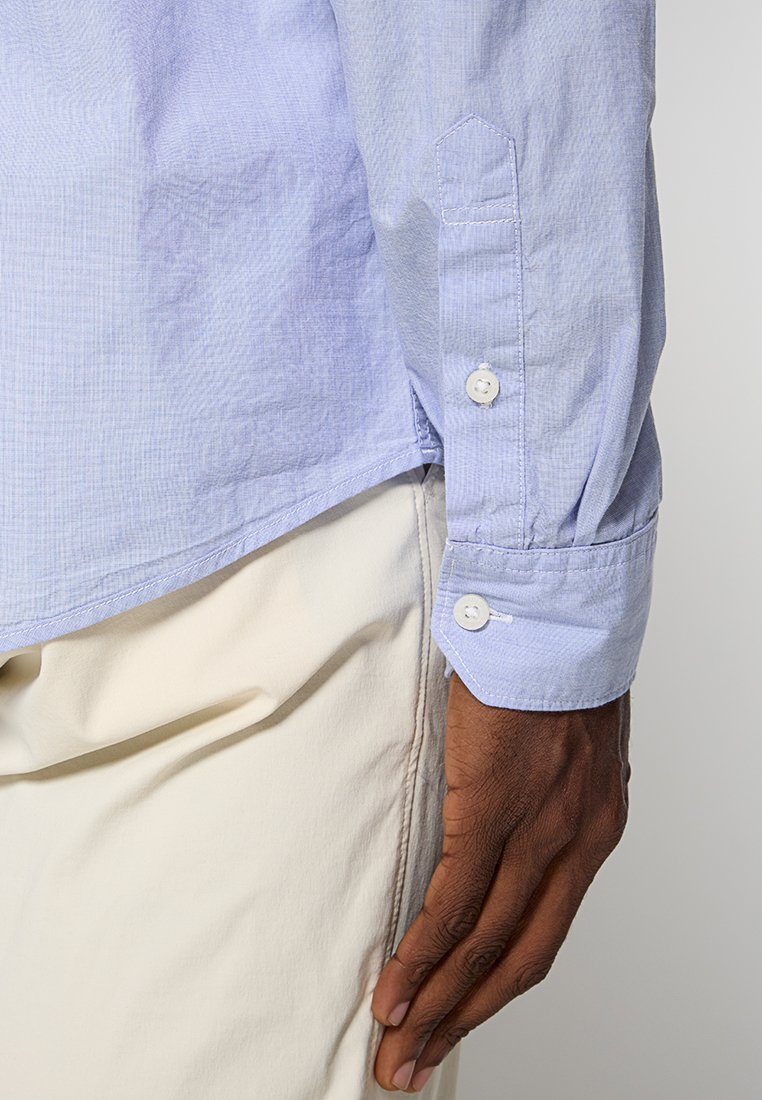 Camicia azzurro chiaro con un sottile motivo a quadretti, dotata di polsini arrotondati e due bottoni bianchi per la chiusura. Pantaloni parzialmente visibili.