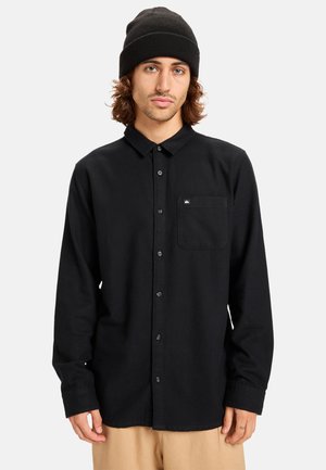Black long-sleeve button-up shirt made of cotton, featuring a chest pocket, adjustable cuffs, and a logo tag on the pocket. Complemented by a black beanie.