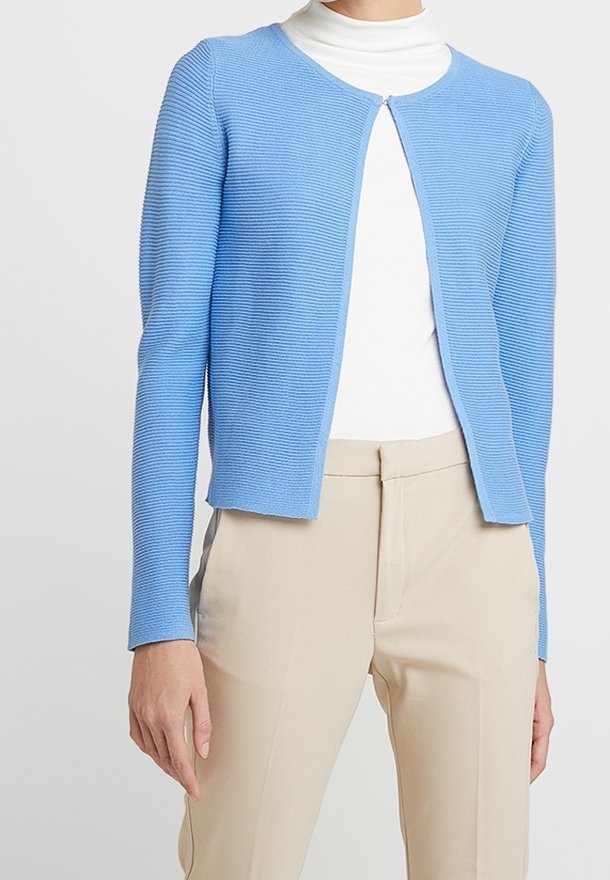 Femme portant un cardigan zippé côtelé bleu clair sur un col roulé blanc, associé à un pantalon beige sur mesure.