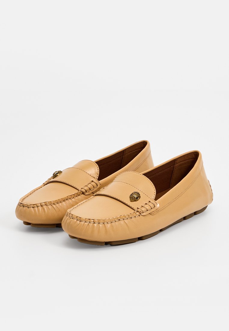 Une paire de mocassins en cuir tan avec des coutures froncées et un petit emblème en laiton sur la sangle, présentée sur un fond blanc.