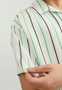 Osoba regulująca krótki rękaw białej koszuli w pionowe zielone i czarne paski, odsłaniająca kołnierz i część szyi.
