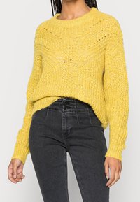 Person in einem strukturierten gelben Strickpullover, der in eine hochtaillierte schwarze Jeans mit Knopfdetails gesteckt ist.