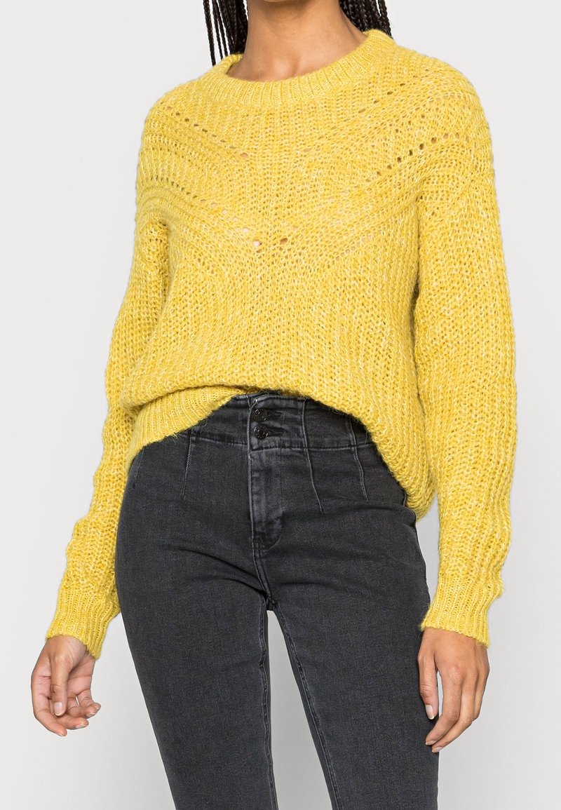Person in einem strukturierten gelben Strickpullover, der in eine hochtaillierte schwarze Jeans mit Knopfdetails gesteckt ist.
