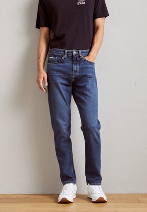 Mørkeblå denim jeans med rett kutt-design, synlig søm, fem-lomme stil og glidelås. Brukt sammen med en svart skjorte og hvite sneakers.
