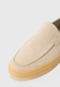 Zapato deslizante de ante beige con detalle de costura en la parte superior y suela de goma marrón claro, mostrado desde un ángulo cercano sobre un fondo blanco.