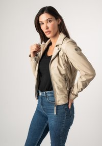 Beige leren motorjack met ritszakken en schouderpatch, gedragen over een zwart topje en blauwe jeans, tegen een effen achtergrond.