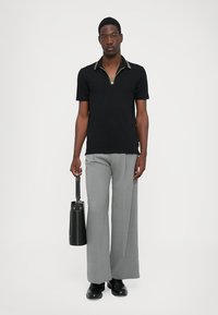 Camisa polo negra con cuello con cremallera, pantalones anchos grises y bolsa negra. Textura suave, diseño minimalista y acentos de cuello en contraste.
