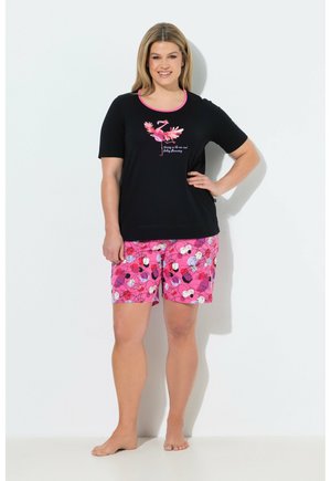 Uśmiechnięta kobieta stoi boso, ubrana w czarną koszulkę z różowym flamingiem i różowe szorty w kwiatowy wzór na białym tle.