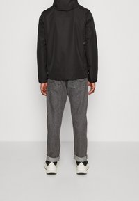 Zwarte waterdichte jas met capuchon, gecombineerd met grijze taps toelopende jeans en witte sneakers, met opgerolde zoom en contrasterend stiksel.