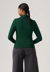 Groen top met lange mouwen, hoge hals en een aansluitende pasvorm, gemaakt van een glad materiaal, gecombineerd met losse grijze broeken met achterzakken.