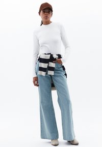 Weißes Langarmshirt, hellblaue weite Jeans und ein gestreifter Schal, der auf der Taille gebunden ist. Eine braune Schirmmütze und beiges Knöchel-Stiefel runden das Outfit ab.