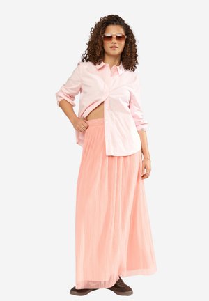 Femme aux cheveux bouclés portant des lunettes de soleil roses, une chemise rose clair à boutons, une longue jupe plissée pêche et des chaussures marron, se tenant avec assurance.