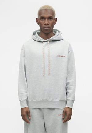 Mladý muž s krátkymi blond vlasmi nosí svetlosivú mikinu s kapucňou a zodpovedajúce tepláky, oba s malým červeným textom „No Problemo“.