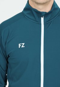 Man som bär en jacka i turkos sportstil med vit vertikal dragkedja och "FZ"-logotyp på vänster bröst mot vit bakgrund.