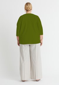 Olivgrünes Oberteil mit ¾ Ärmeln, mit geradem Schnitt und Rückennaht, kombiniert mit hellbeigen weiten Hosen. Glatte Textur, minimaler Detail.