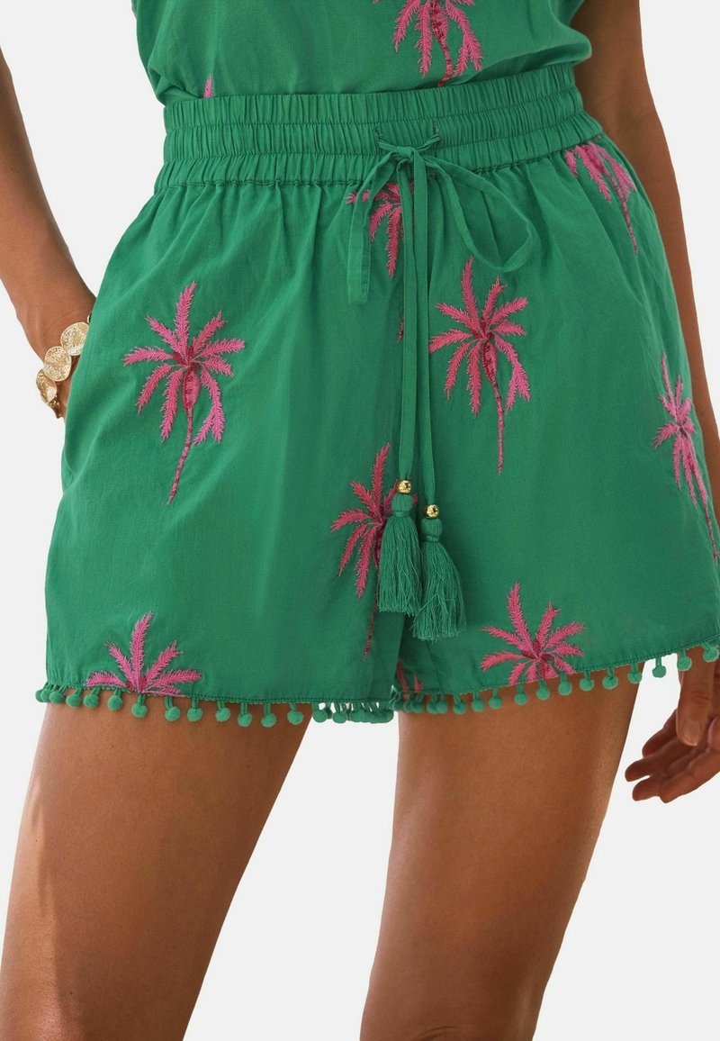 Grüne Shorts mit rosa Palmenmuster, elastischem Bund mit Kordelzug und Quasten, Pompom-Borte am Saum, getragen mit einem goldenen Armband.