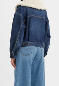 Farkkutakki tekoturkiskauluksella, kahdella sivutaskulla ja napilletuilla hihansuiden resoreilla. Päällä sinisten leveälahkeisten farkkujen päällä, takakuvassa.