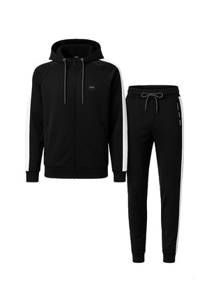 Svart träningsoverall med en zip-up hoodie med vita sidostråkar och matchande byxor med dragsko i midjan. Tillverkad av mjukt, stretchigt material.