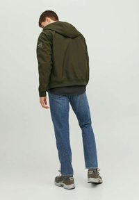 Olive green hooded jacket with a relaxed fit, elastic cuffs, and waistband, paired with blue jeans and gray sneakers.