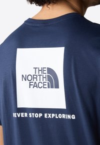 Námořnická modrá bavlněná trička s bílým čtvercovým logem na zadní straně, pod nímž je text "THE NORTH FACE" a "NEVER STOP EXPLORING".