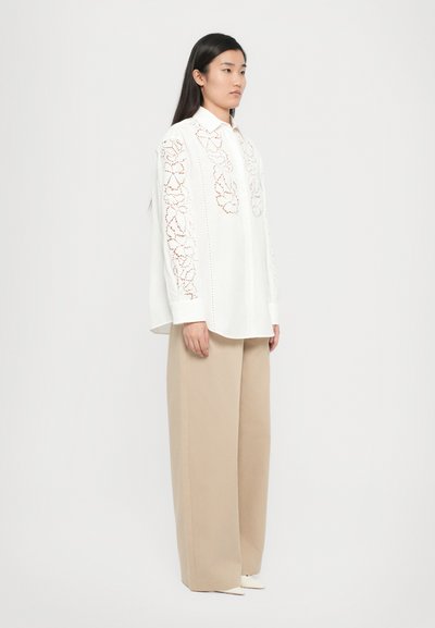 Camisa de algodón blanca con bordado floral a lo largo de las mangas, combinada con pantalones beige de corte holgado y zapatos blancos de punta.