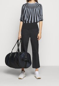 Czarna i jasnoniebieska w paski bluzka z okrągłym dekoltem, w zestawieniu z czarnymi spodniami 3/4 i białymi trampkami, trzymająca dużą czarną torbę podróżną.
