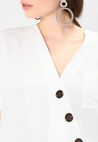 Hvid v-hals bluse med korte ærmer, der har en lomme i ikke og fire brune rundknapper langs venstre side. Store runde rhinestones øreringe.