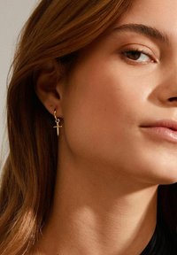 Goldene Creolenohrringe mit einem kleinen Kreuzanhänger. Die Ohrringe sind glänzend und haben eine glatte Oberfläche, sie hängen unterhalb des Ohrläppchens.
