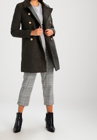 Mørkegrøn uldblandingsfrakke med guldknapper, grå sweater, sort-hvid ternede bukser og sorte ankelstøvler.