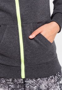 Sudadera gris con cremallera amarilla, con bolsillos frontales y puños acanalados. Tejido texturizado con un tacto suave.