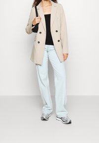 Kvinna som bär en beige dubbelknäppt kavaj, svart topp, ljusblå raka jeans, silverfärgade sneakers och bär en liten svart axelväska.