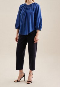 Blusa azzurra a maniche lunghe con scollatura arricciata e polsi elasticizzati, abbinata a pantaloni neri a tre quarti e tacchi neri con cinturini. Sfondo neutro.