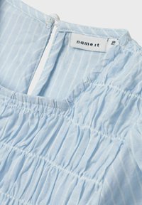 Top azzurro chiaro a maniche corte con righe bianche verticali. Presenta una texture arricciata e una scollatura rotonda con chiusura sul retro. Etichetta del marchio visibile.