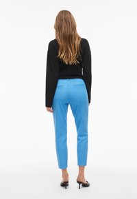 Mustern mustrine pikkade varrukatega must kampsun, millele on sobitatud helesinised kitsaste lõikega püksid ja mustad avatud varvastega kontsad; vaade tagant.