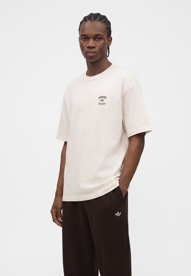 Ung mand med flettet hår iført en beige Adidas t-shirt og mørke Adidas bukser, stående med den ene hånd i lommen.